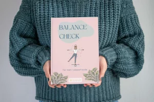 Hände halten das Heft „Balance-Check“ vor einem grauen Strickpullover