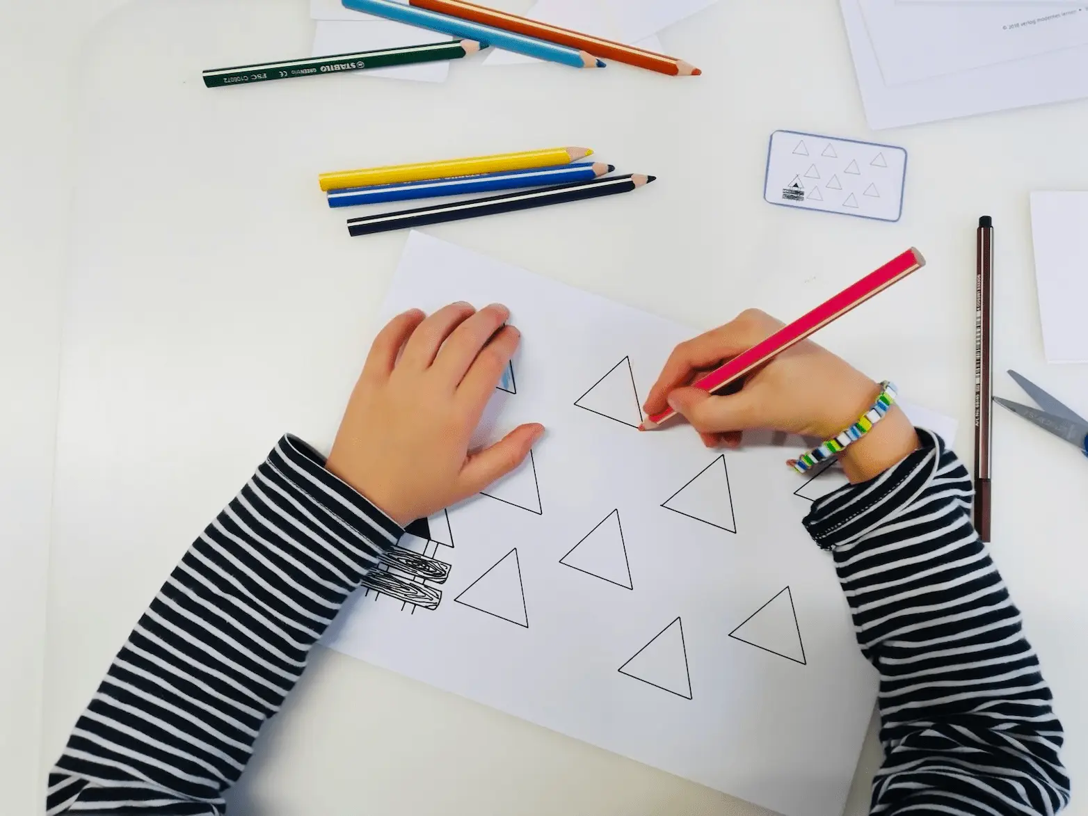 Kinderhände malen mit Bleistift Dreiecke nach, daneben Buntstifte